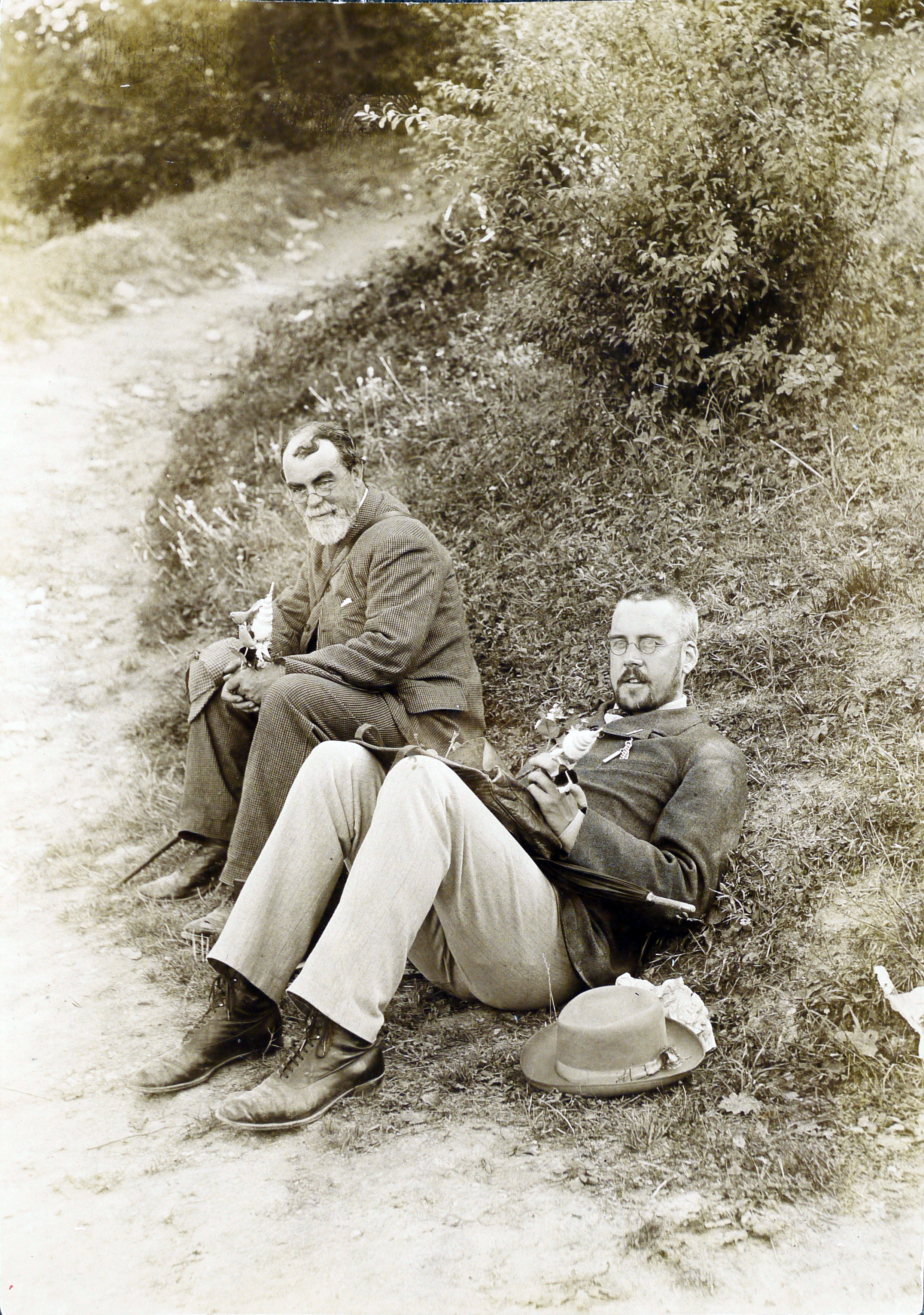 A black-and-white photograph by Alfred Cathie, depicting Samuel Butler and Henry Festing Jones sitting on grass by a path. Both wear walking boots and hold flowers. Butler's hat is balanced on his knee, Festing Jones's rested on the ground; Festing Jones holds an umbrella on his lap.