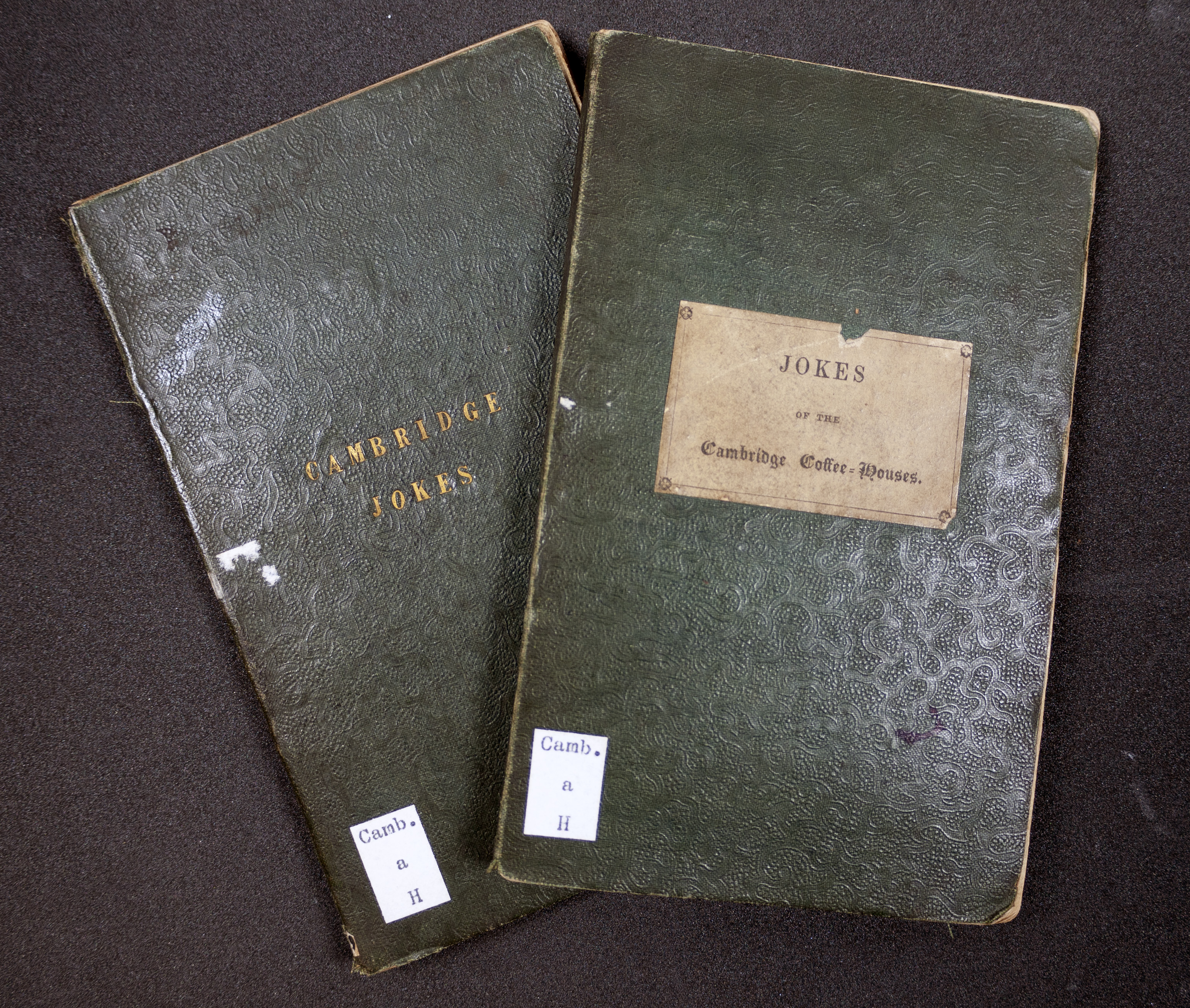 Two small green-bound books, overlaid so that the titles on their covers remain visible. The left- and bottommost book has 'Cambridge Jokes' embossed in gold, and the right- and topmost bears a paper label: 'Jokes of the Cambridge Coffee-Houses'.
